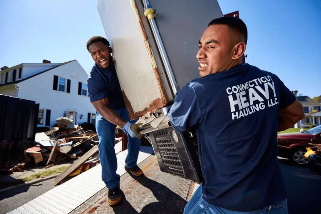 Two professional junk removal workers loading heavy item into trailer on residential property in Connecticut